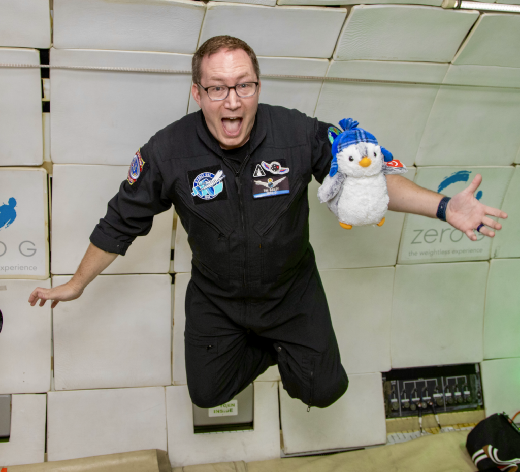 Tim Bailey floating in a stunning and form fitting black flight suit with an adorable stuffed penguin floating in front of him.